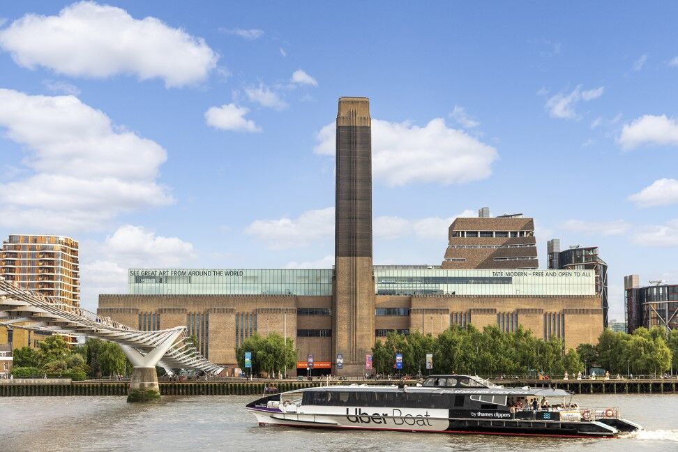 An Uber Boat crosses under the Millennium Bridge on the River Thames. In the background the Tate Modern is visible.