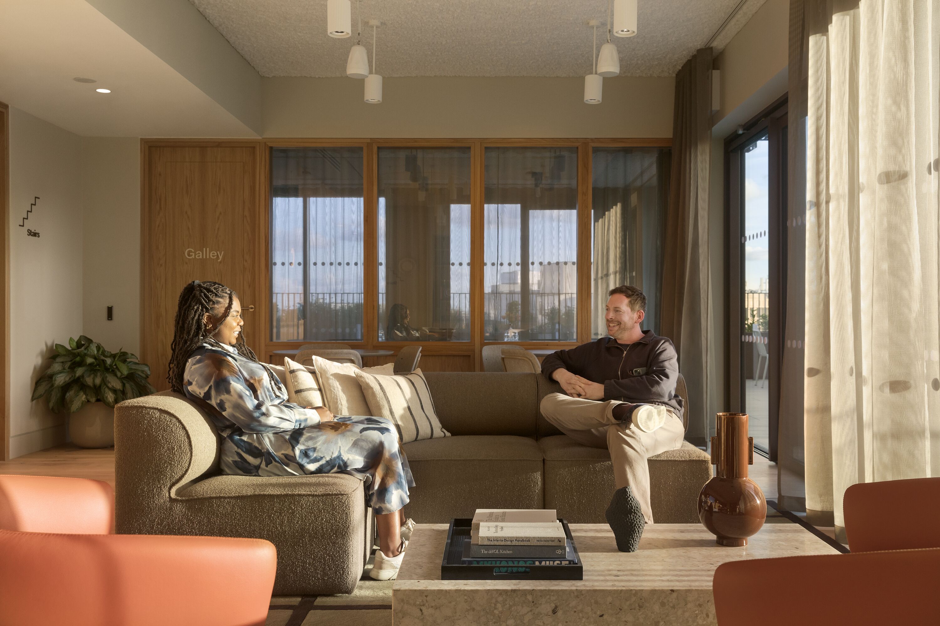 Two people having a relaxed conversation in a cozy, sunlit lounge area with comfortable sofas. The room has large windows, wooden accents, and contemporary decor elements.